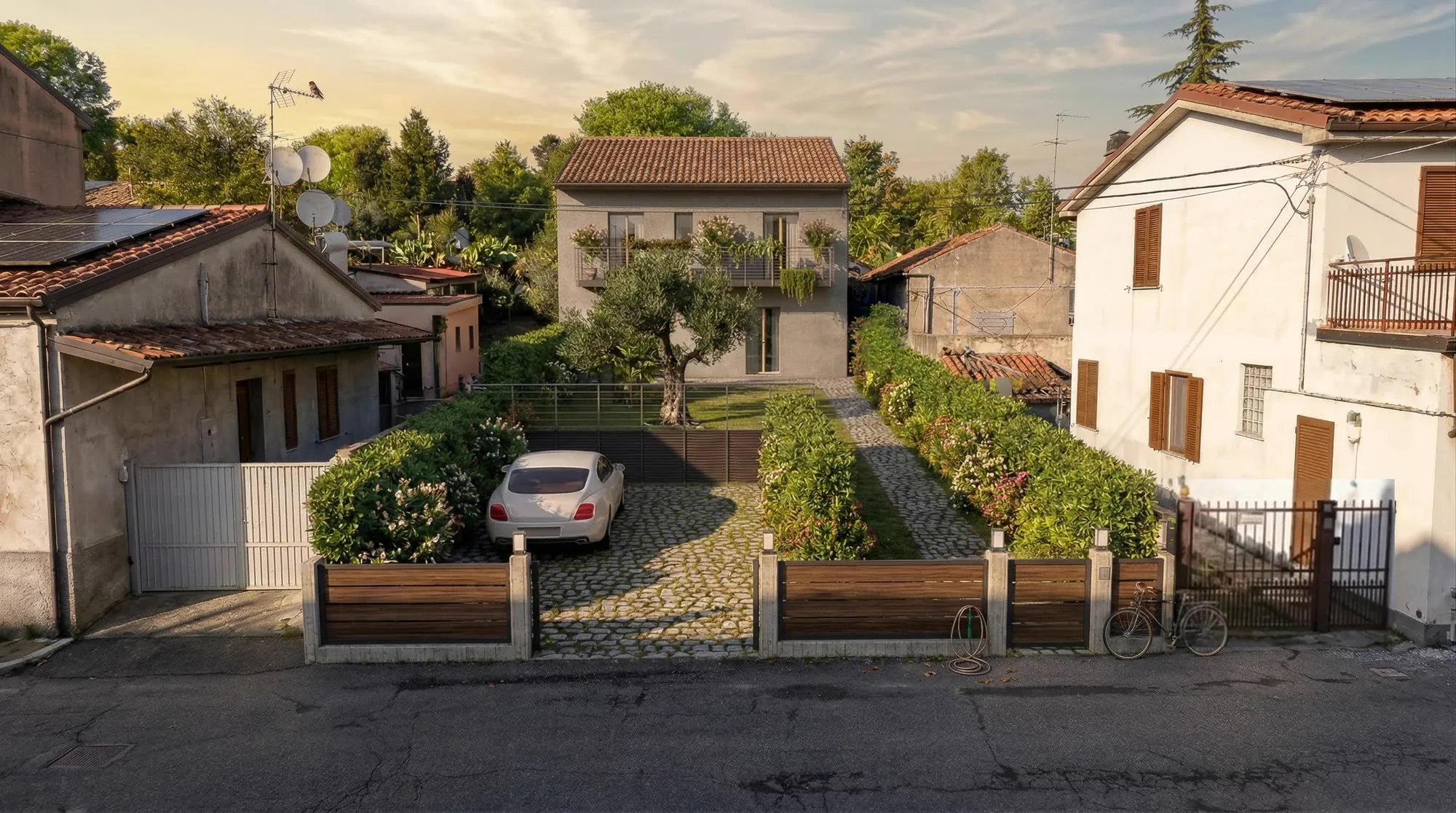 Casa a due piani vista dalla strada al tramonto: giardino con ulivo, vialetto in pietra, auto nel cortile e recinzione in legno