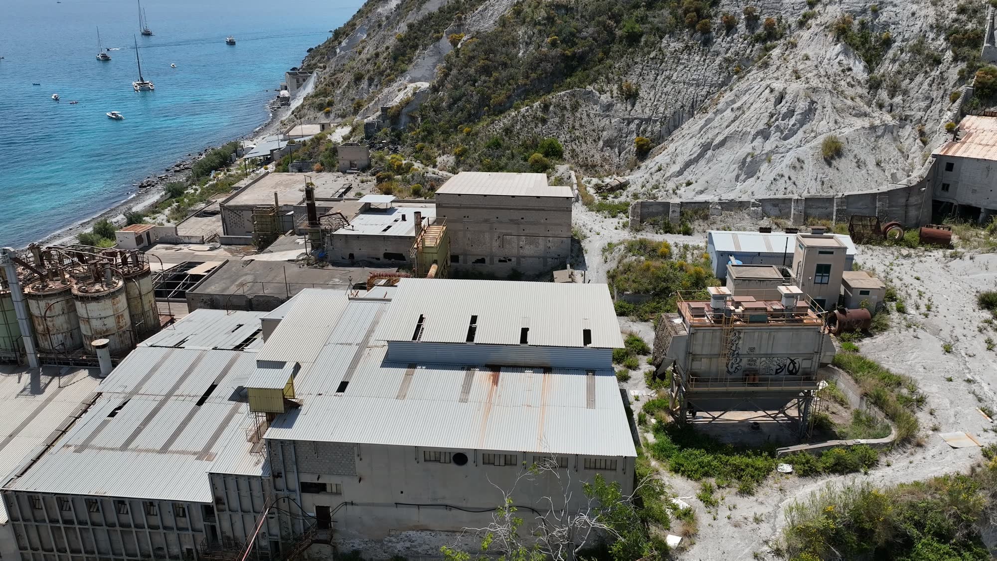 Complesso industriale dismesso sulla costa visto dal drone: capannoni, silos e il versante chiaro alle spalle
